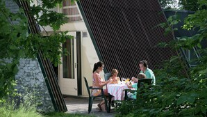 Une terrasse avec une table et des chaises, entourée de verdure et bordée d’un bâtiment doté d’un toit en pente.