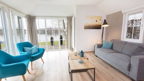 A modern living room with a grey sofa, two blue armchairs, a wooden coffee table, and a view of a lake through large windows. - Stylish Woodland Chalet (Hoge Hexel)