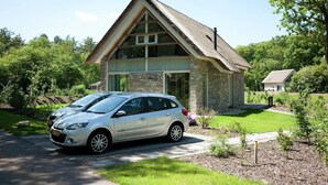 Une petite maison de plain-pied, dotée d’un toit de chaume et de grandes fenêtres, entourée d’un jardin bien entretenu et avec une voiture garée à proximité.