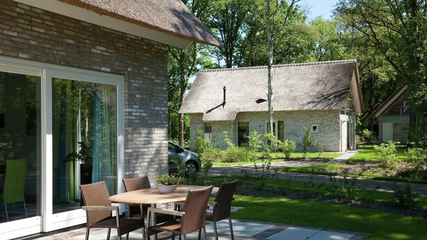 Une maison moderne avec une façade en briques, un toit de chaume et une terrasse équipée d’une table et de chaises.