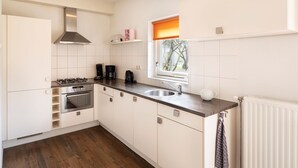 Una cocina moderna con armarios blancos, una campana extractora de acero inoxidable, estufa a gas, horno doble, un fregadero y una ventana con vistas a la vegetación.