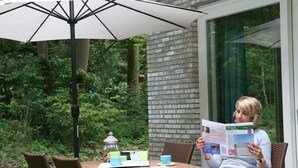 Eine Person liest eine Zeitschrift an einem Tisch, der von einem weißen Sonnenschirm beschattet wird, umgeben von Korbstühlen und einem Glasbehälter.