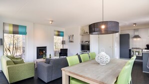 Ein modernes Wohnzimmer mit Kamin, eine Essecke mit Holztisch und grünen Stühlen sowie eine Küche mit grauen Schränken und einem Gaskochfeld.