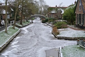 Ein verschneiter Weg führt zu einer Brücke über einen Kanal, umgeben von Wohnhäusern und viel Grün.