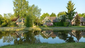 A tranquil lakeside setting with three houses, each with distinct architectural styles, surrounded by lush greenery and a calm pond reflecting the buildings. - Thatched Villa in National Park (Paasloo)