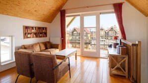 A cozy loft-style room with a wooden ceiling, a large window with red curtains, a sofa, a table, and a bookshelf.