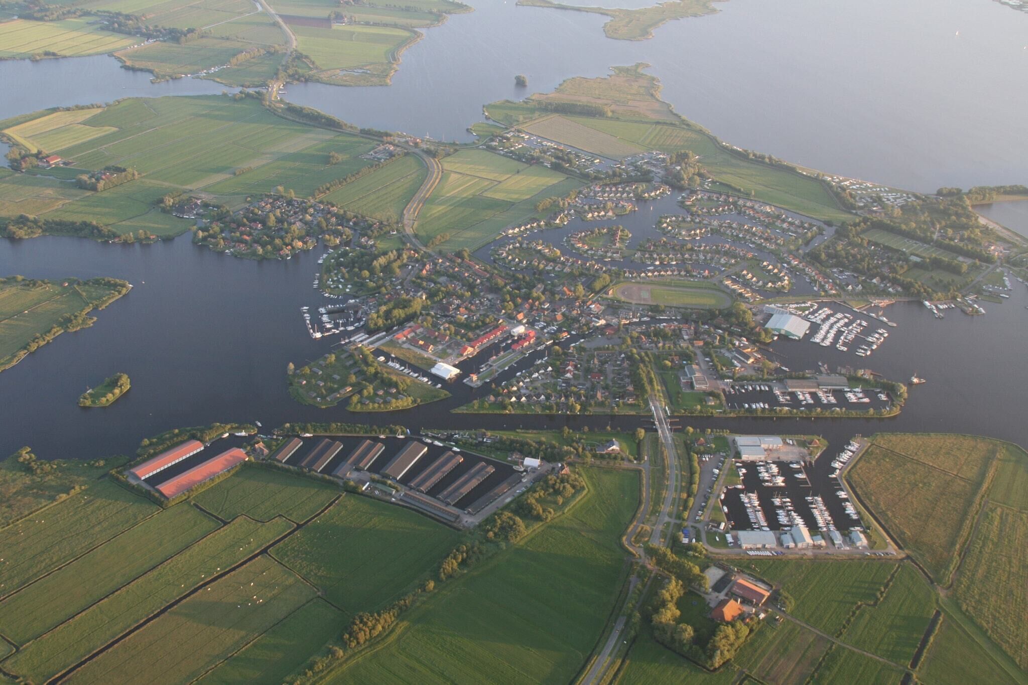 An aerial view of a coastal town with a marina, green fields, and industrial buildings.
