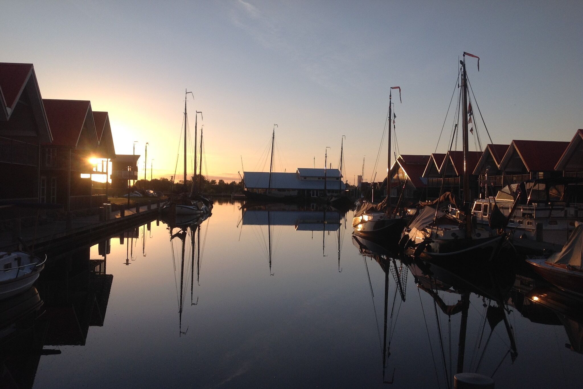 A marina with boats docked, a calm water surface reflecting the buildings, and a sunset in the background.