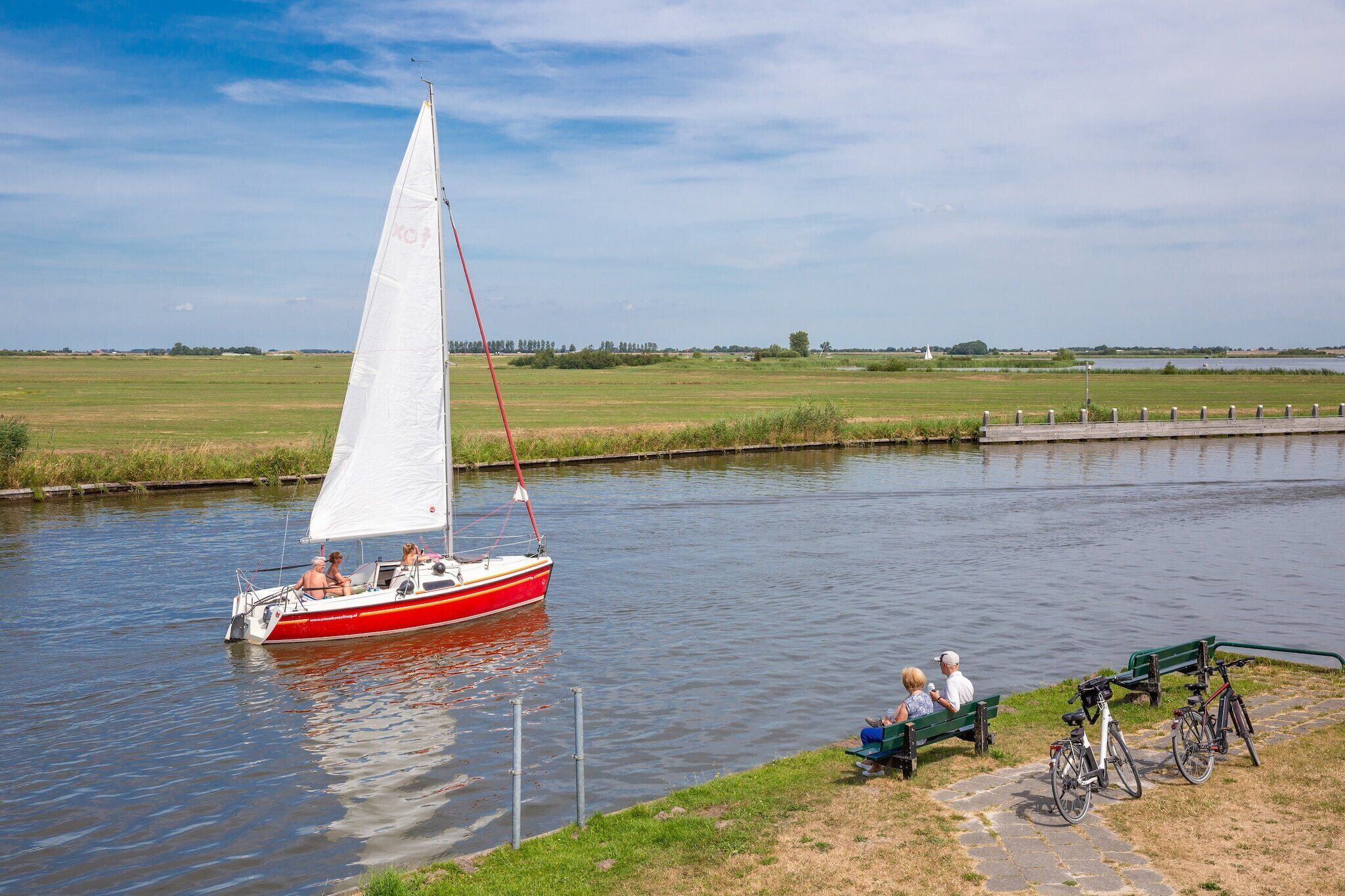 Een zeilboot op een rustige rivier met een grasrijke oever en een pad.