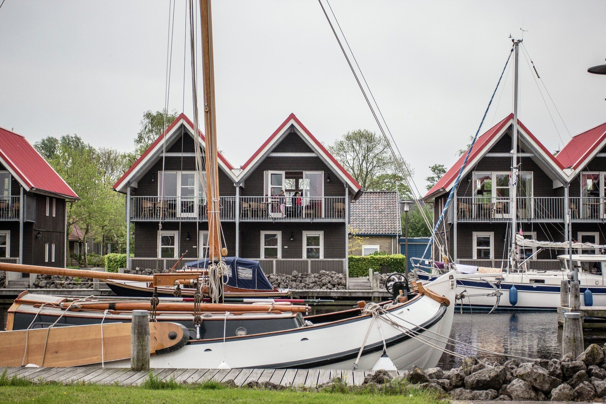 Un port de plaisance où sont amarrés plusieurs bateaux, des embarcations en bois avec des mâts, et des maisons aux toits rouges et aux balcons.