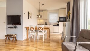 Un salon moderne doté d’un téléviseur à écran plat, d’un coin repas avec une table en bois et des chaises blanches, d’une cuisine avec des meubles blancs et un four à micro-ondes, et d’une grande fenêtre agrémentée de rideaux.