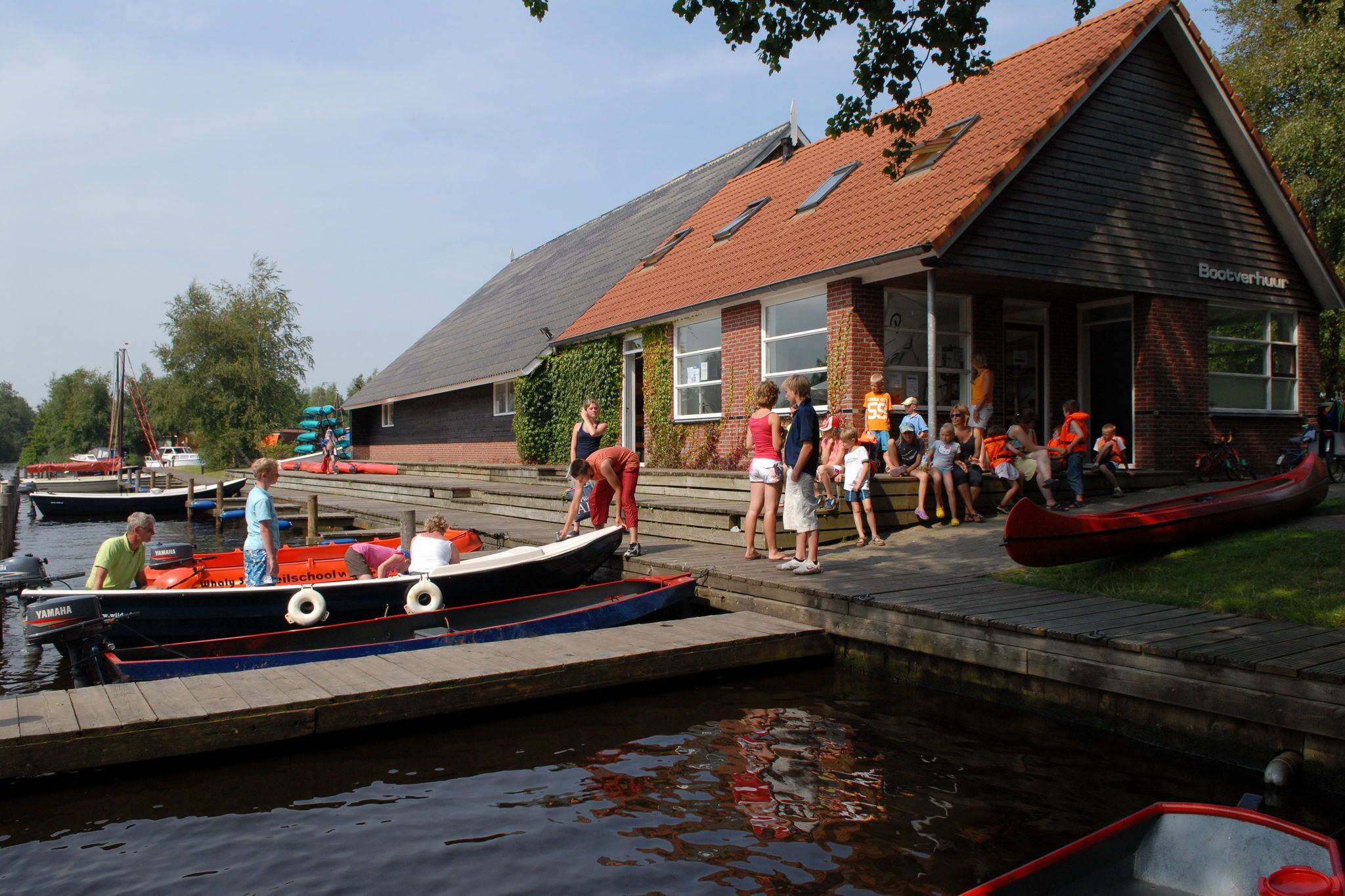 A docked boat with people boarding, a building with a red roof, and a calm water body.