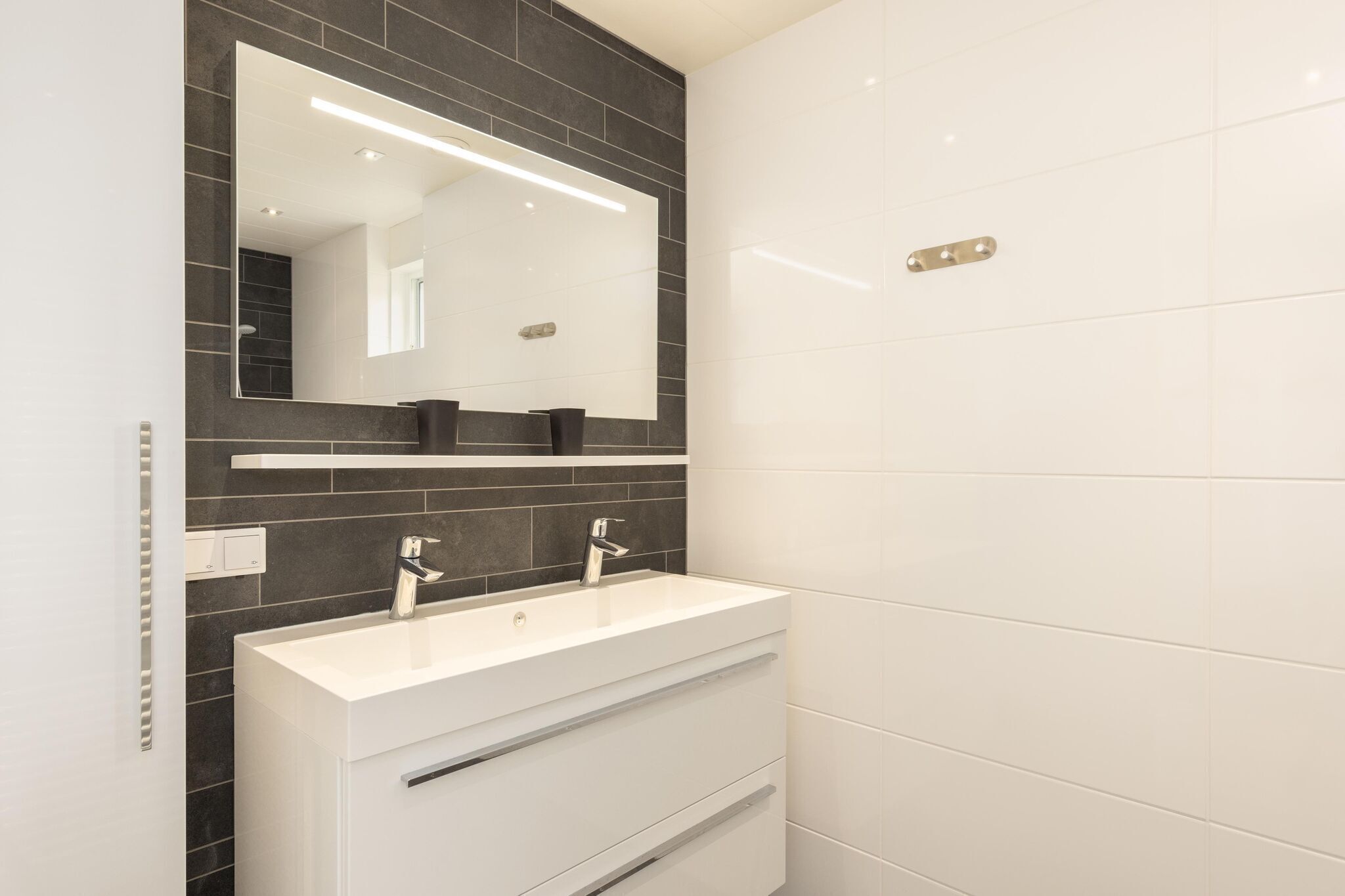 A modern bathroom with a white vanity, dark tiled walls, and a large mirror.