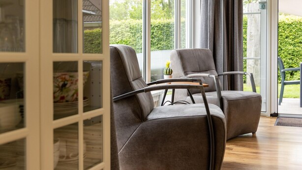 Une pièce moderne avec deux fauteuils gris, une petite table sur laquelle est posée une plante en pot, et de grandes fenêtres donnant sur un paysage verdoyant.