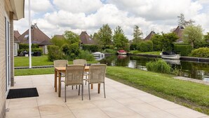 A patio with a table and chairs, a grassy area, and a canal with boats. - Cozy Bungalow with Terrace (Eernewoud)