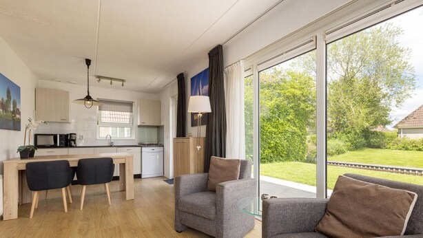 A modern kitchen with a dining area, two grey armchairs, and a large window with a view of greenery. - Cozy Bungalow with Terrace (Eernewoud)