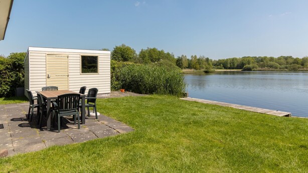 Una cabaña pequeña de un solo nivel con un patio y una mesa con sillas, ubicada cerca de un lago con una zona verde y árboles al fondo.