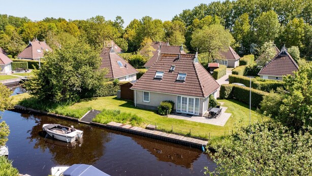 A canal-side property with a house featuring a tiled roof, a small boat docked by the water, and lush greenery surrounding the area. - Nature Bungalow with Two Baths (Eernewoud)