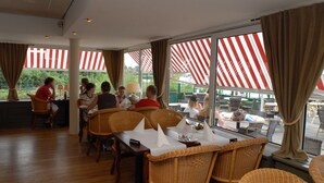 A restaurant with wicker chairs and tables, large windows with red and white striped awnings, and a view of greenery outside. - Nature Bungalow with Two Baths (Eernewoud)