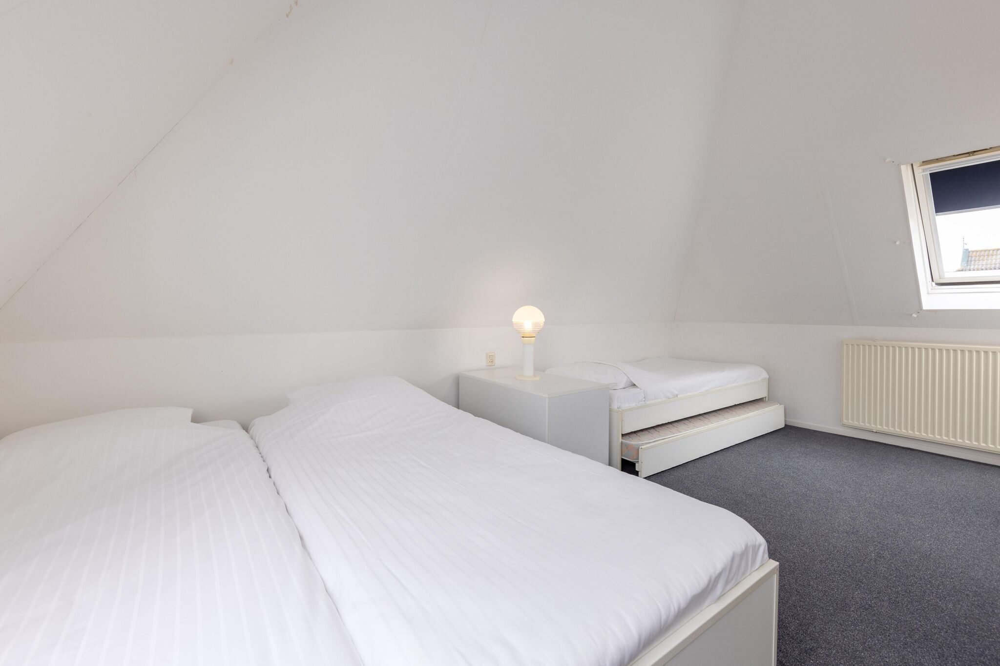 Een kleine, witte kamer met eenpersoonsbedden, een nachtkastje met een lamp, een raam en een radiator.