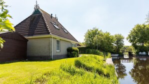 A house with a sloped roof, a small garden, and a canal in the background. - Nature Bungalow with Two Baths (Eernewoud)