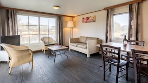 Ett hotellrum med en soffa, ett matbord med stolar, en TV och ett fönster med gardiner.