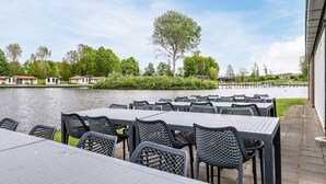 Ett utomhusområde vid floden med bord och stolar, fri sikt över floden och en rad byggnader i bakgrunden.