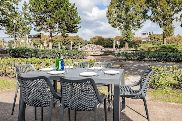 Un espace de restauration en plein air avec une table dressée pour six personnes, entourée de chaises, dans un cadre verdoyant évoquant un parc, avec des arbres et un mur de pierre en arrière-plan.
