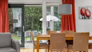 A dining area with a wooden table and wicker chairs, a grey sofa, and a large window with red curtains. - Forest Stay with Shared Pool (Borger)