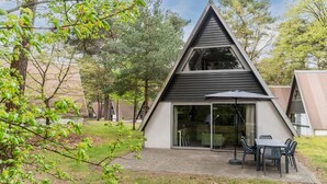 A modern house with a triangular roof, a patio with a table and chairs, and a glass door leading to a room.