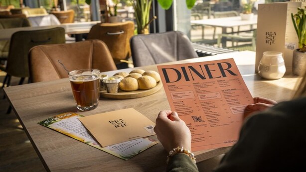 A person holding a menu at a restaurant table with a drink and a plate of food.