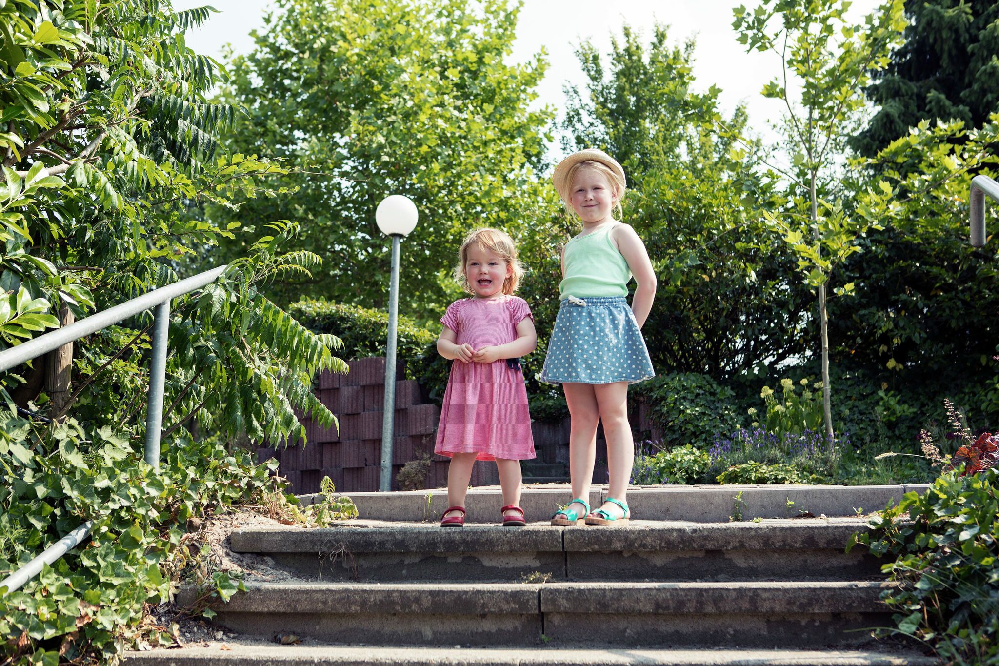 Two children standing on outdoor steps surrounded by greenery.