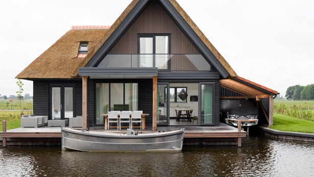 Et moderne hus med stråtækt tag, store vinduer og en båd i havnen foran.