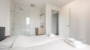 Et moderne hotelværelse med en stor seng, et bruseområde med glasvægge, et trækommode og et spejl.