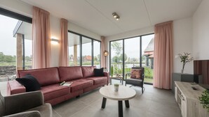 En moderne stue med en rød sofa, et rundt bord og udsigt til det fri gennem store vinduer.