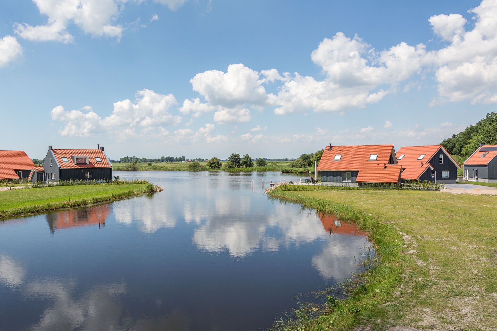 En lugn miljö vid floden, med moderna hus som har röda tak och stora fönster, omgivna av frodig grönska och en klar himmel.