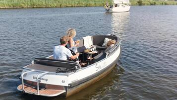 Een smalle motorboot met twee personen aan boord, een klein tafeltje en een zeilboot op de achtergrond.