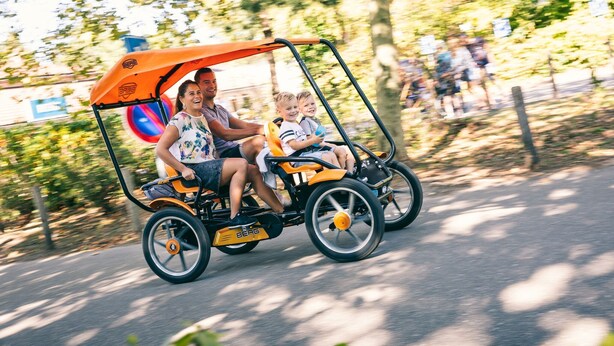 A family in a small, open-air vehicle with a canopy, driving on a paved road surrounded by trees. - Designer Chalet in the Veluwe (Wissel/Epe)