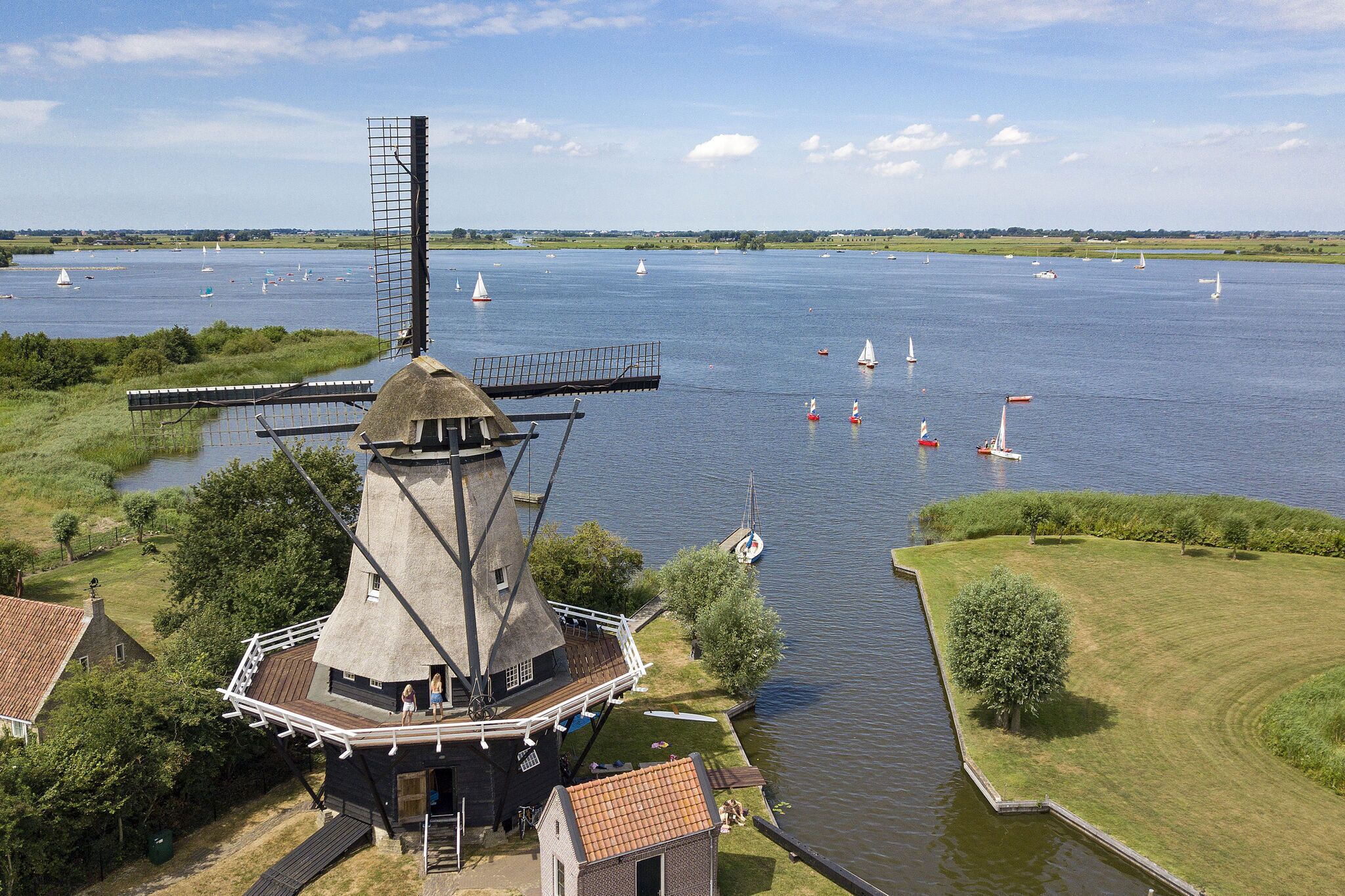 Eine traditionelle Windmühle mit Aussichtsplattform, umgeben von einem ruhigen Fluss mit Segelbooten und einem gepflegten Rasen.