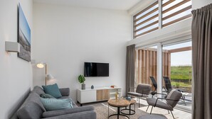 A modern living room with a sofa, a coffee table, and a TV mounted on the wall. There is a window with curtains and a view of greenery outside.