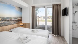 A hotel room with two beds, a large window with curtains, a TV, and a shower area.
