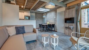 Een moderne woonkamer met een bank, twee kleine krukjes, een eettafel met stoelen, een aan de muur bevestigde televisie en een groot raam met gordijnen.
