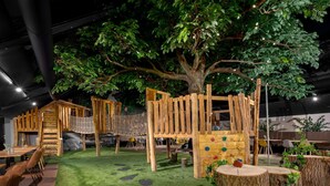 Un espace de jeux pour enfants avec un grand arbre, des structures d’escalade en bois et une surface de jeu souple.