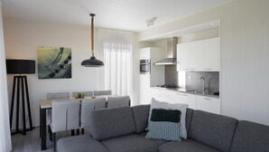 A modern living room with a grey sofa, a dining table with chairs, a pendant light, and a kitchen area with white cabinets and appliances.