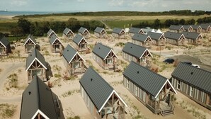 A series of wooden cabins with pitched roofs, arranged in rows on sandy terrain with a view of the sea and distant fields.