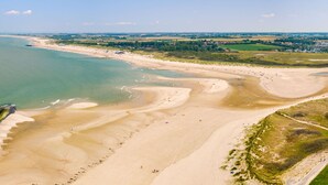 An aerial view of a beach with clear waters, sandy shores, and a mix of open fields and buildings in the background.