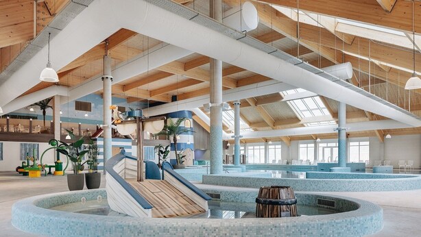 A spacious indoor swimming pool area with a wooden ceiling, large windows, and a central slide.