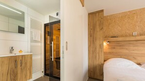 A modern hotel room with a wooden headboard, a white bed, a wooden vanity with a mirror, and a glass door leading to a bathroom. - Wellness Stay with Sauna (Borger)