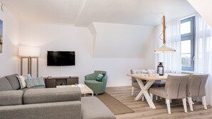 Een moderne woonkamer met een grijze bank, een groene fauteuil, een flatscreen televisie, een eettafel met stoelen en een groot raam met gordijnen.