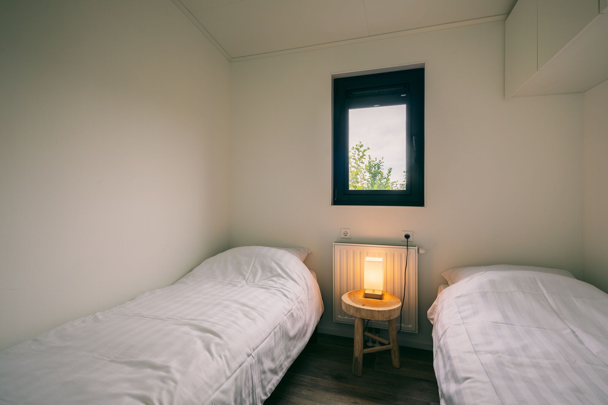 A small, single-room accommodation with two single beds, a small table with a lamp, and a window with a view of greenery.